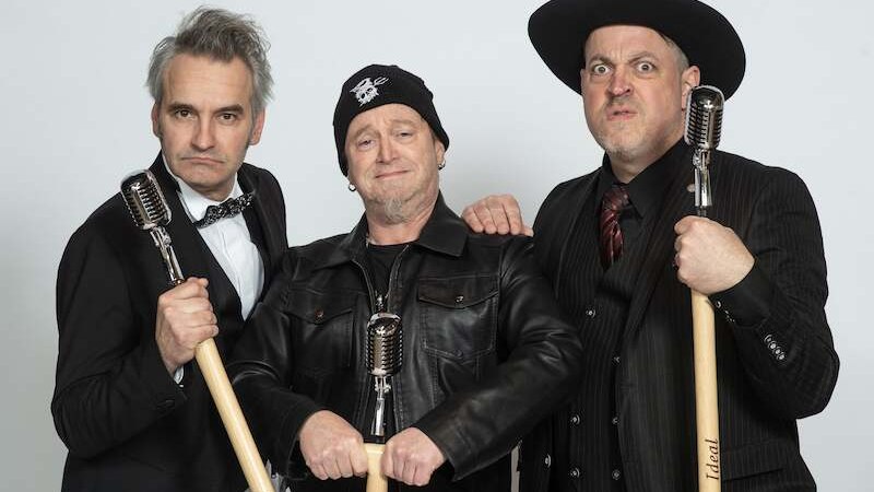 Drei M&auml;nner posieren mit Mikrofonen. Einer tr&auml;gt eine Fliege, der andere einen Hut, der dritte eine M&uuml;tze. Sie wirken humorvoll und selbstbewusst., &copy; C&sup2; CONCERTS GmbH