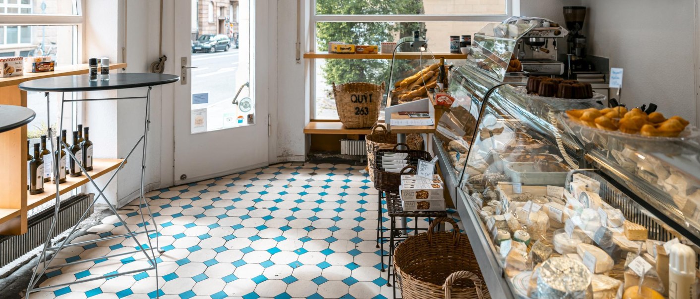 Ein Feinkostladen mit Käseauslage, Gebäck und Weinflaschen. Helle Fliesen und ein Fenster mit Blick auf die Straße., © SMG, Sarah Schmid Ein Feinkostladen mit Käseauslage, Gebäck und Weinflaschen. Helle Fliesen und ein Fenster mit Blick auf die Straße., © SMG, Sarah Schmid