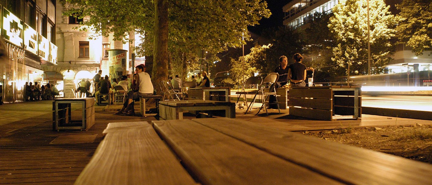 Nachtaufnahme der Theodor-Heuss-Straße in Stuttgart. Menschen sitzen auf Holzbänken unter Bäumen, umgeben von beleuchteten Gebäuden und Straßenlaternen., © Stuttgart-Marketing GmbH, Marc Gilardone