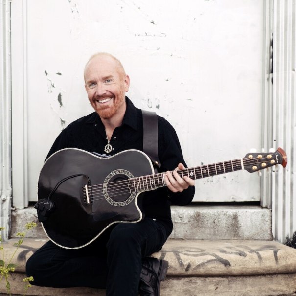 Ein Mann mit rotem Bart sitzt lächelnd auf einer Treppe und hält eine schwarze Akustikgitarre. Der Hintergrund ist eine weiße Wand., © Mauerwerk GmbH Ein Mann mit rotem Bart sitzt lächelnd auf einer Treppe und hält eine schwarze Akustikgitarre. Der Hintergrund ist eine weiße Wand., © Mauerwerk GmbH