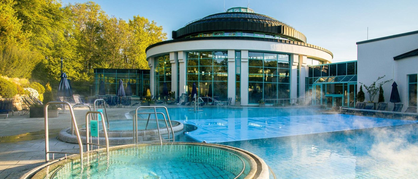 Die Mineraltherme Böblingen zeigt einen dampfenden Außenpool vor einem modernen, runden Gebäude mit großen Glasfenstern., © Stuttgart-Marketing GmbH, Sarah Schmid