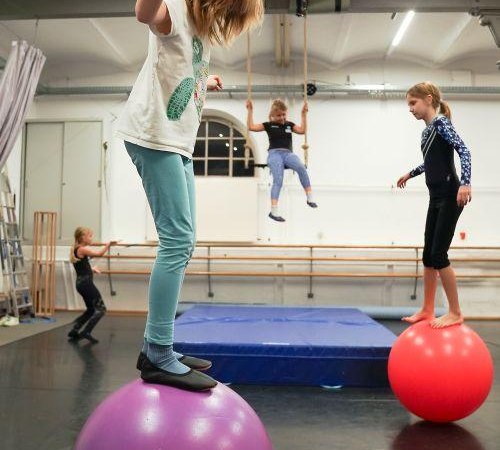 Kinder balancieren auf bunten Gymnastikb&auml;llen in einem Zirkustrainingsraum. Im Hintergrund schwingt ein Kind an einem Trapez., &copy; Kunstschule Labyrinth