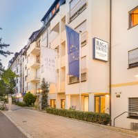 Außenansicht des Rega Hotels in Stuttgart. Mehrstöckiges Gebäude mit Fahnen und beleuchteten Straßenlaternen. Ruhige Straße mit geparkten Autos., © TOMAS