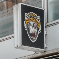 Ein Schild mit dem Logo von Kap Tormentoso hängt an einem modernen Gebäude. Es zeigt ein Segelschiff und dekorative Elemente., © Stuttgart Marketing GmbH, Sarah Schmid