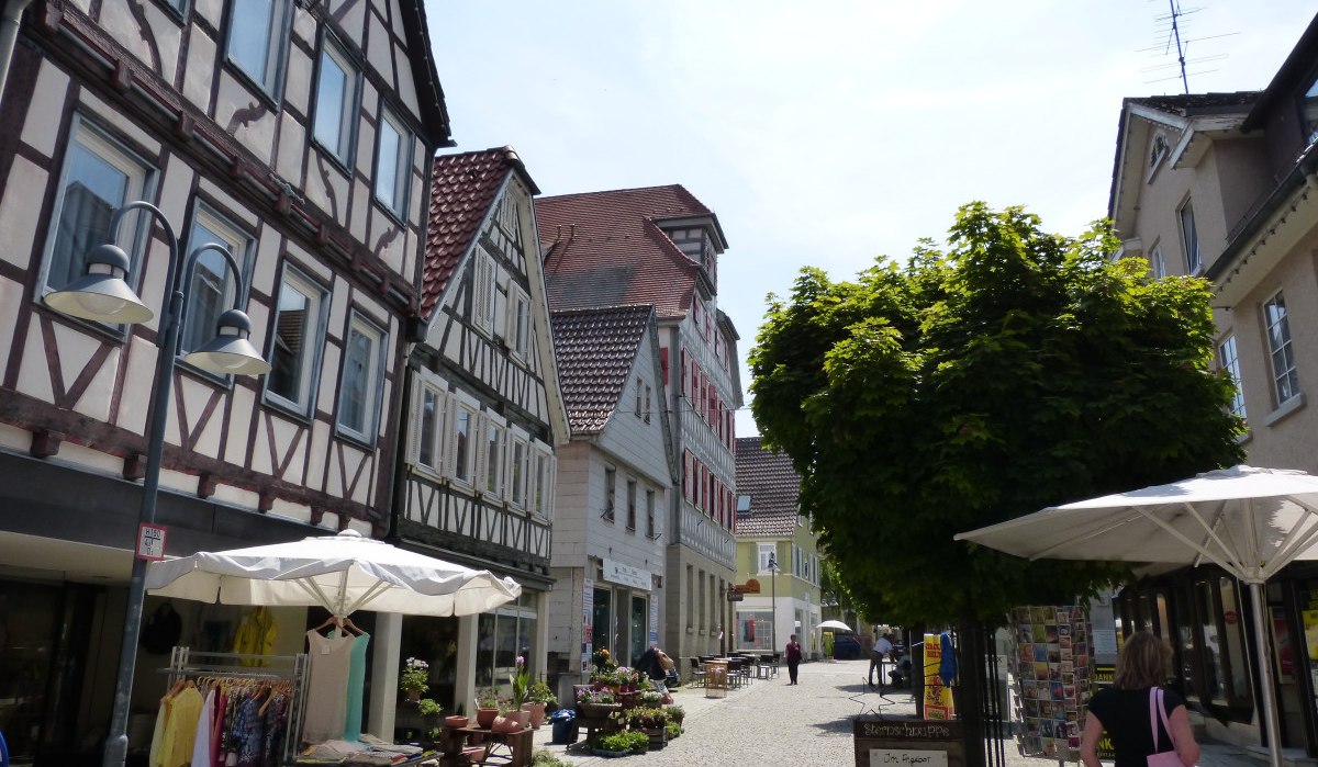 Fußgängerzone in Vaihingen an der Enz mit Fachwerkhäusern, Geschäften und Passanten. Sonnenschirme und ein Baum spenden Schatten., © Land der 1000 Hügel - Kraichgau-Stromberg Fußgängerzone in Vaihingen an der Enz mit Fachwerkhäusern, Geschäften und Passanten. Sonnenschirme und ein Baum spenden Schatten., © Land der 1000 Hügel - Kraichgau-Stromberg