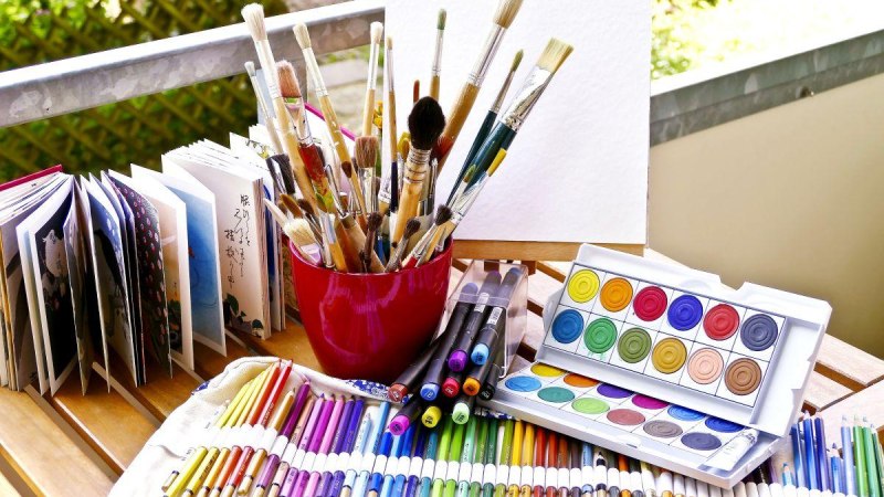 Ein Tisch mit Malutensilien: Pinsel in einem roten Becher, Buntstifte, Filzstifte, Wasserfarben und ein aufgeschlagenes Buch., © © Astrid Untermann Ein Tisch mit Malutensilien: Pinsel in einem roten Becher, Buntstifte, Filzstifte, Wasserfarben und ein aufgeschlagenes Buch., © © Astrid Untermann