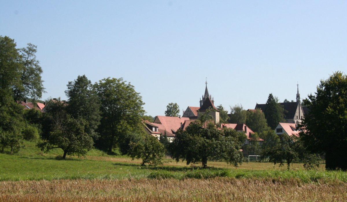 Blick auf Bebenhausen mit roten Dächern und Kirchturm, umgeben von grünen Bäumen und Feldern unter klarem Himmel., © Natur.Nah. Schönbuch & Heckengäu Blick auf Bebenhausen mit roten Dächern und Kirchturm, umgeben von grünen Bäumen und Feldern unter klarem Himmel., © Natur.Nah. Schönbuch & Heckengäu
