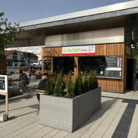 Kleiner Kiosk 'Asia Delight by Doan' mit Holzverkleidung und Sitzbereich im Freien. Menschen sitzen an Tischen, umgeben von Pflanzen., © Asia Delight GmbH