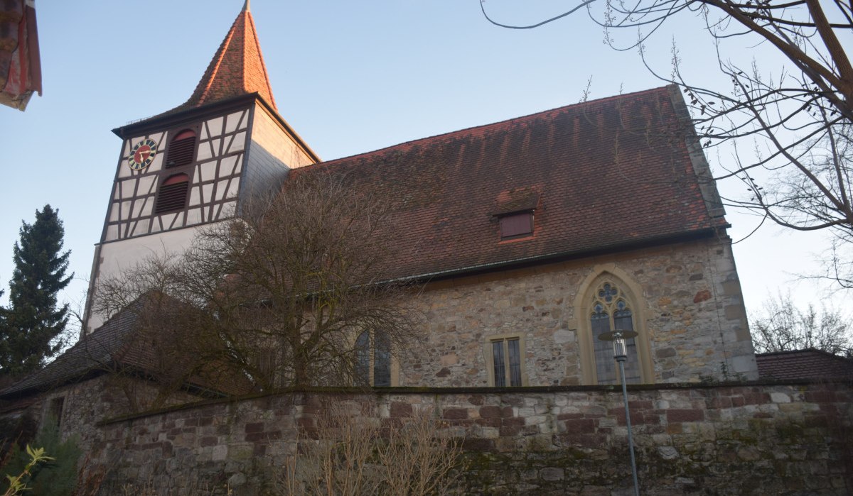 Die Evangelische St. Veitskirche mit Fachwerkturm und rotem Dach vor blauem Himmel, umgeben von Bäumen und einer Steinmauer., © Land der 1000 Hügel - Kraichgau-Stromberg Die Evangelische St. Veitskirche mit Fachwerkturm und rotem Dach vor blauem Himmel, umgeben von Bäumen und einer Steinmauer., © Land der 1000 Hügel - Kraichgau-Stromberg