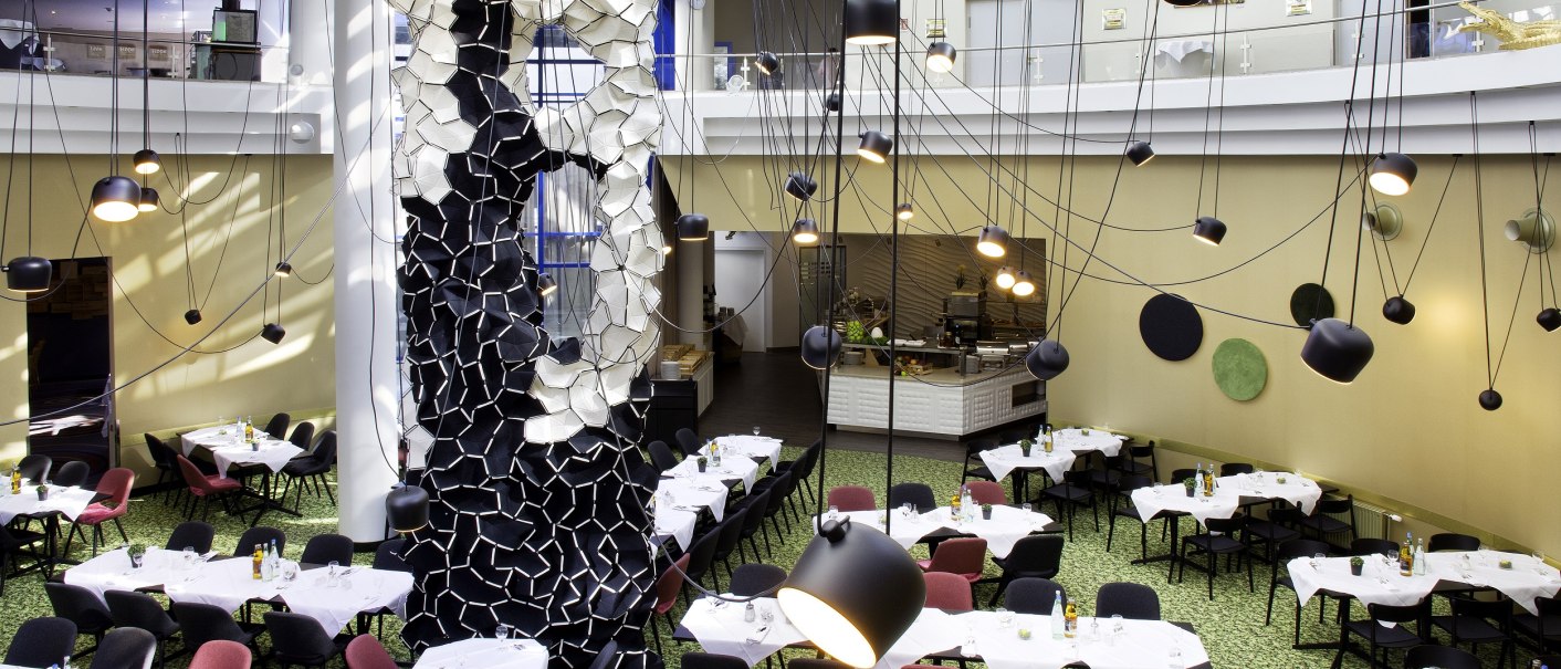 Ein modernes Restaurant mit künstlerischer Dekoration, hängenden Lampen und gedeckten Tischen auf grünem Teppichboden., © Holiday Inn Ein modernes Restaurant mit künstlerischer Dekoration, hängenden Lampen und gedeckten Tischen auf grünem Teppichboden., © Holiday Inn