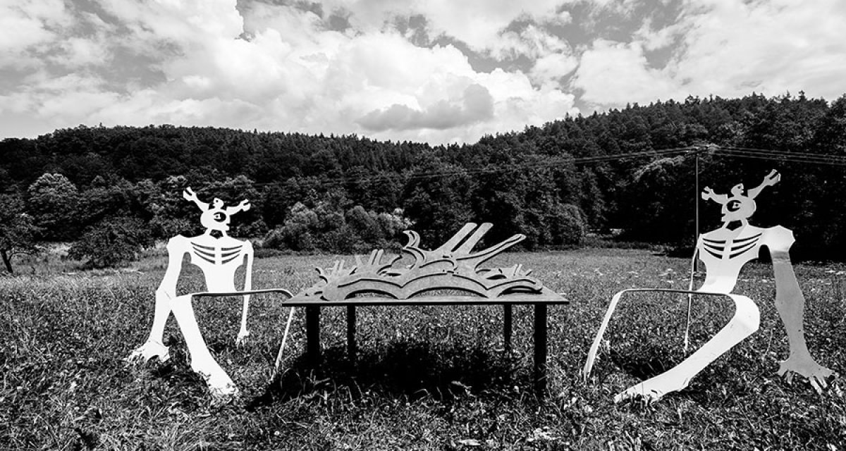 Zwei stilisierte Metallfiguren sitzen auf einer Wiese an einem Tisch mit Buchmotiv. Im Hintergrund erstreckt sich ein Wald unter einem bew&ouml;lkten Himmel., &copy; Natur.Nah. Sch&ouml;nbuch & Heckeng&auml;u