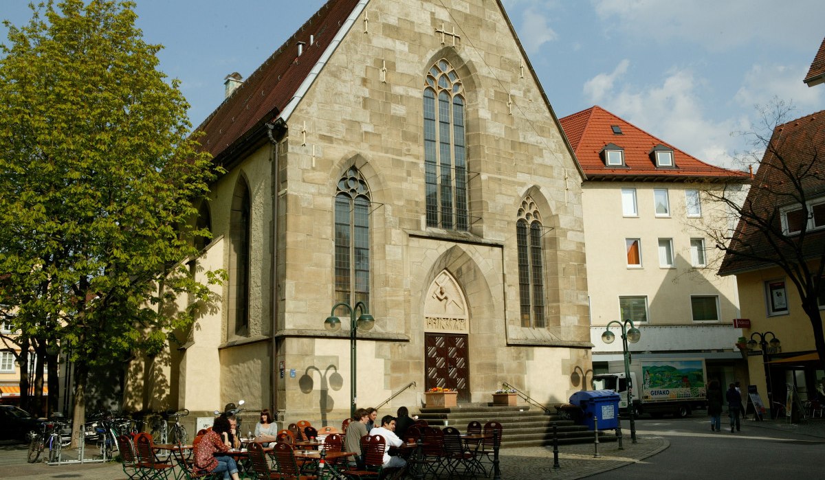 Die Nikolaikirche mit gotischen Fenstern, umgeben von Bäumen und Sitzgelegenheiten. Menschen sitzen draußen bei sonnigem Wetter., © Sphäre Verlag