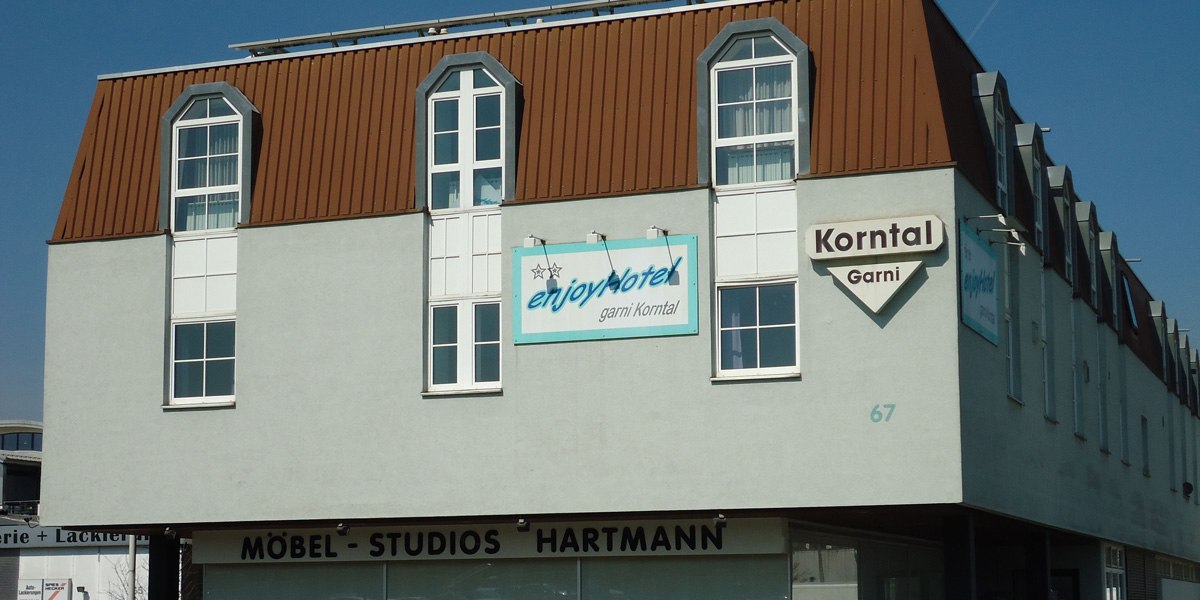 Ein Gebäude mit Schildern von enjoyHotel garni Korntal und Möbel-Studios Hartmann. Das Dach ist braun, die Fassade grau., © enjoyHotel garni Korntal Stuttgart