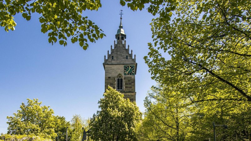 Ein Kirchturm erhebt sich &uuml;ber gr&uuml;ne B&auml;ume unter einem klaren blauen Himmel. Die Szene strahlt Ruhe und Naturverbundenheit aus., &copy; SMG, Sarah Schmid