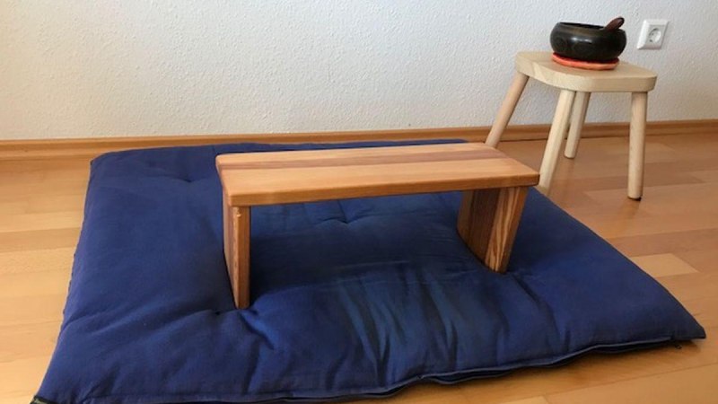 Ein Meditationskissen mit einem Holzhocker darauf, daneben ein kleiner Tisch mit einer Klangschale auf einem Holzboden., © Bürgerengagement und Bürgerhäuser Ein Meditationskissen mit einem Holzhocker darauf, daneben ein kleiner Tisch mit einer Klangschale auf einem Holzboden., © Bürgerengagement und Bürgerhäuser
