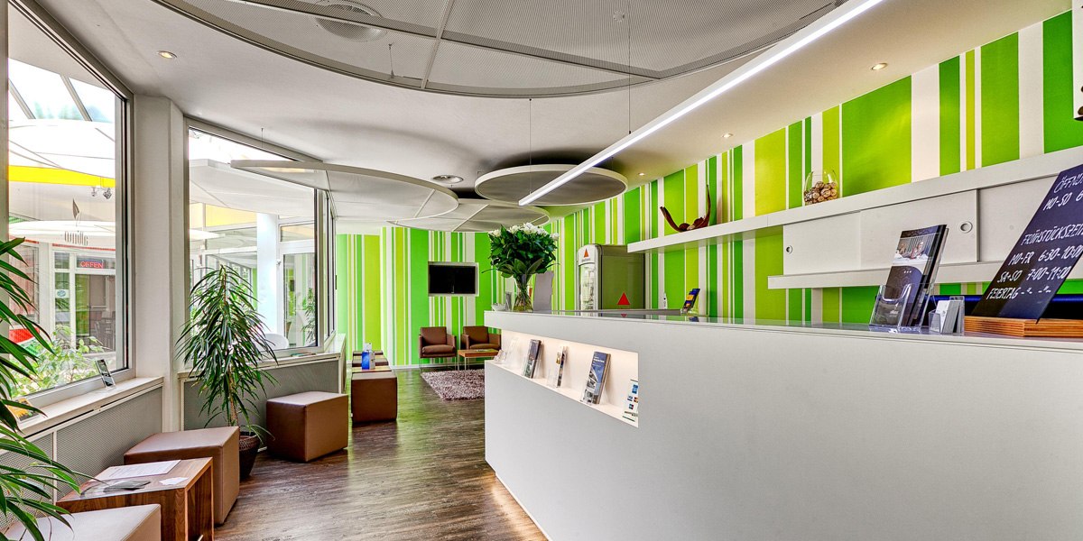 Moderne Rezeption mit grün-weiß gestreifter Wand, Pflanzen und Sitzbereich. Helle, einladende Atmosphäre mit Broschüren und Dekoration., © attimo