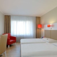 Modernes Hotelzimmer mit zwei Einzelbetten, rotem Sessel, Schreibtisch und Fenster mit Vorhängen. Rote Lampen sorgen für Akzente., © Hotel Stuttgart-Sindelfingen City by Tulip Inn