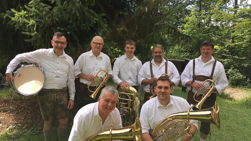 Eine Blaskapelle in traditioneller Kleidung posiert mit Instrumenten im Freien vor Bäumen. Die Musiker tragen weiße Hemden und Lederhosen., © Alb 7 Eine Blaskapelle in traditioneller Kleidung posiert mit Instrumenten im Freien vor Bäumen. Die Musiker tragen weiße Hemden und Lederhosen., © Alb 7