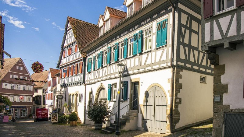 Fachwerkhäuser mit blauen Fensterläden in einer sonnigen Altstadtstraße, dekoriert mit Blumen. Kopfsteinpflaster und klare Himmel im Hintergrund., © Stuttgart-Marketing GmbH, Sarah Schmid Fachwerkhäuser mit blauen Fensterläden in einer sonnigen Altstadtstraße, dekoriert mit Blumen. Kopfsteinpflaster und klare Himmel im Hintergrund., © Stuttgart-Marketing GmbH, Sarah Schmid