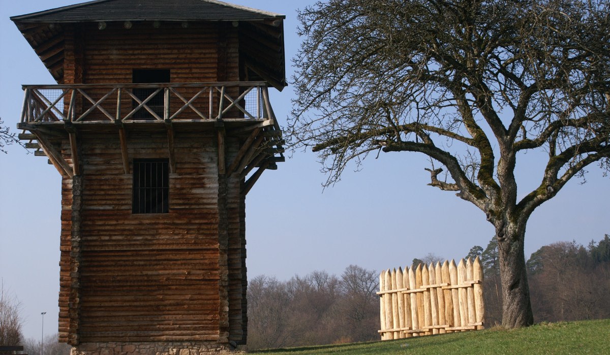 Ein hölzerner römischer Wachturm steht neben einem kahlen Baum und einem kleinen Holzzaun auf einer grünen Wiese., © Remstal Tourismus e.V.