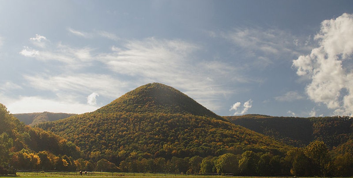 Ein runder, bewaldeter Berg erhebt sich unter einem blauen Himmel mit Wolken. Im Vordergrund sind grüne Felder und Bäume zu sehen., © Bad Urach Tourismus