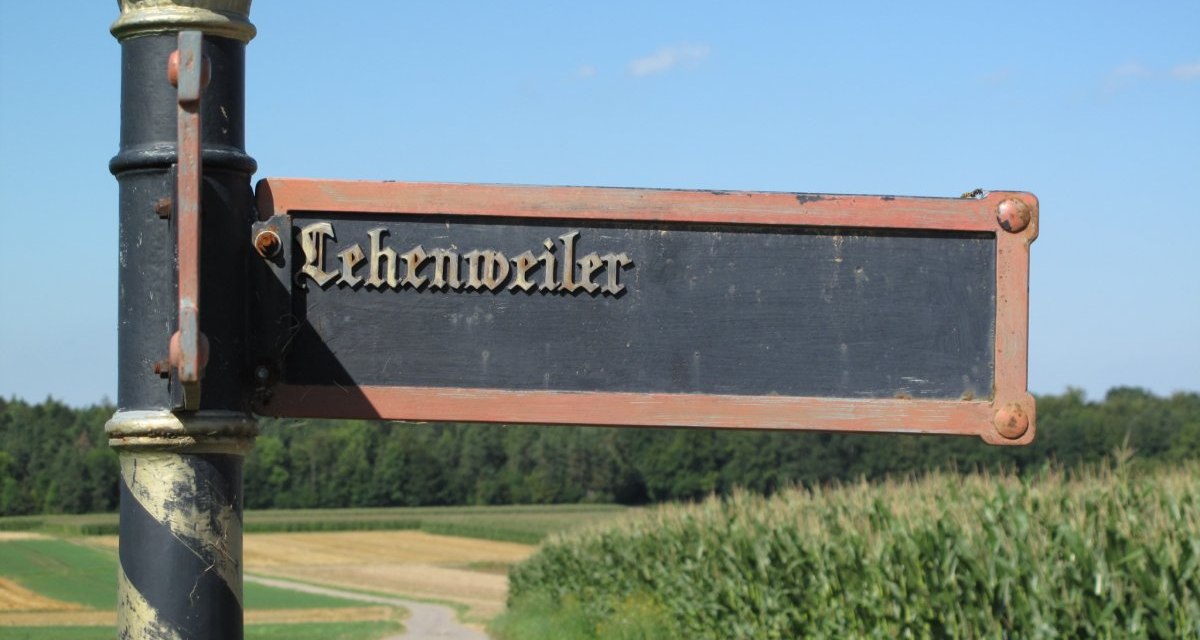 Ein altes Wegweiser-Schild mit der Aufschrift 'Lehenweiler' steht vor einem Feld und Wald unter klarem Himmel., © Natur.Nah. Schönbuch & Heckengäu Ein altes Wegweiser-Schild mit der Aufschrift 'Lehenweiler' steht vor einem Feld und Wald unter klarem Himmel., © Natur.Nah. Schönbuch & Heckengäu
