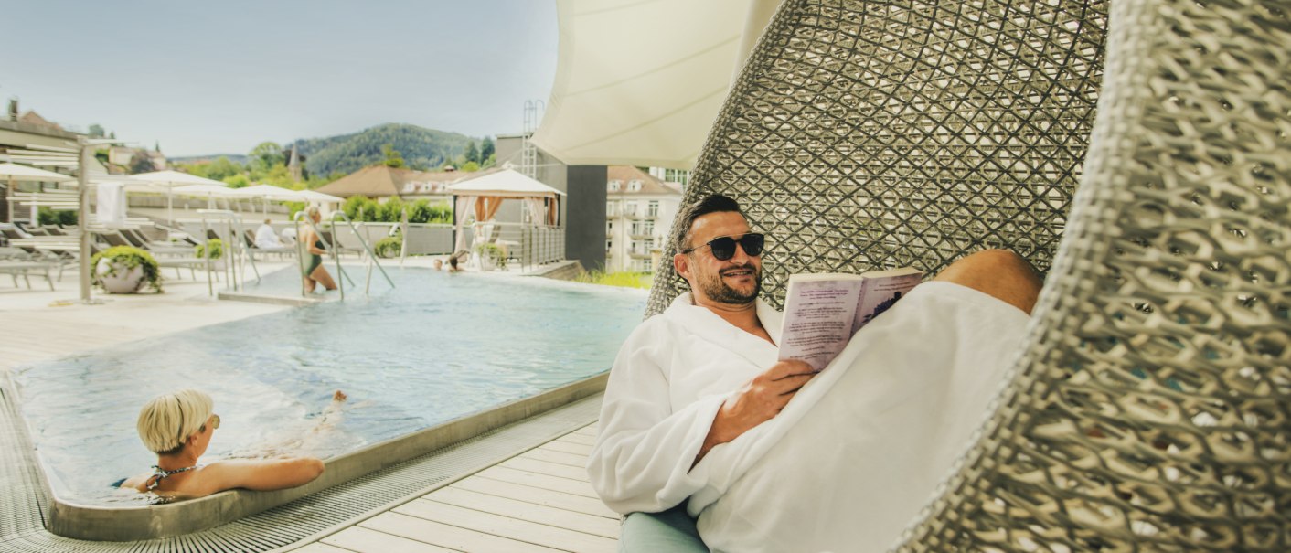 Ein Mann entspannt in einem Bademantel auf einer Liege am Pool des Palais Thermal. Im Hintergrund genie&szlig;en weitere G&auml;ste das Wasser und die Sonne., &copy; &copy;LOCHER Fotodesign & Manufaktur &copy;Staatsbad Wildbad GmbH