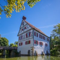 Fachwerkhaus mit roten Fensterläden an einem Teich, umgeben von Bäumen und einer Brücke im Hintergrund, unter blauem Himmel., © SMG, Achim Mende