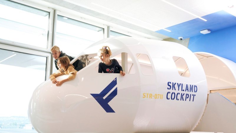 Drei Kinder spielen in einem Cockpit-Nachbau mit der Aufschrift 'SkyLand Cockpit' und blicken aus den Fenstern. Helle, moderne Umgebung., © Flughafen Stuttgart Drei Kinder spielen in einem Cockpit-Nachbau mit der Aufschrift 'SkyLand Cockpit' und blicken aus den Fenstern. Helle, moderne Umgebung., © Flughafen Stuttgart