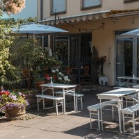 Gemütlicher Außenbereich eines Cafés mit weißen Tischen und Stühlen, umgeben von üppigen Pflanzen und bunten Blumen., © Stuttgart Marketing GmbH, Sarah Schmid