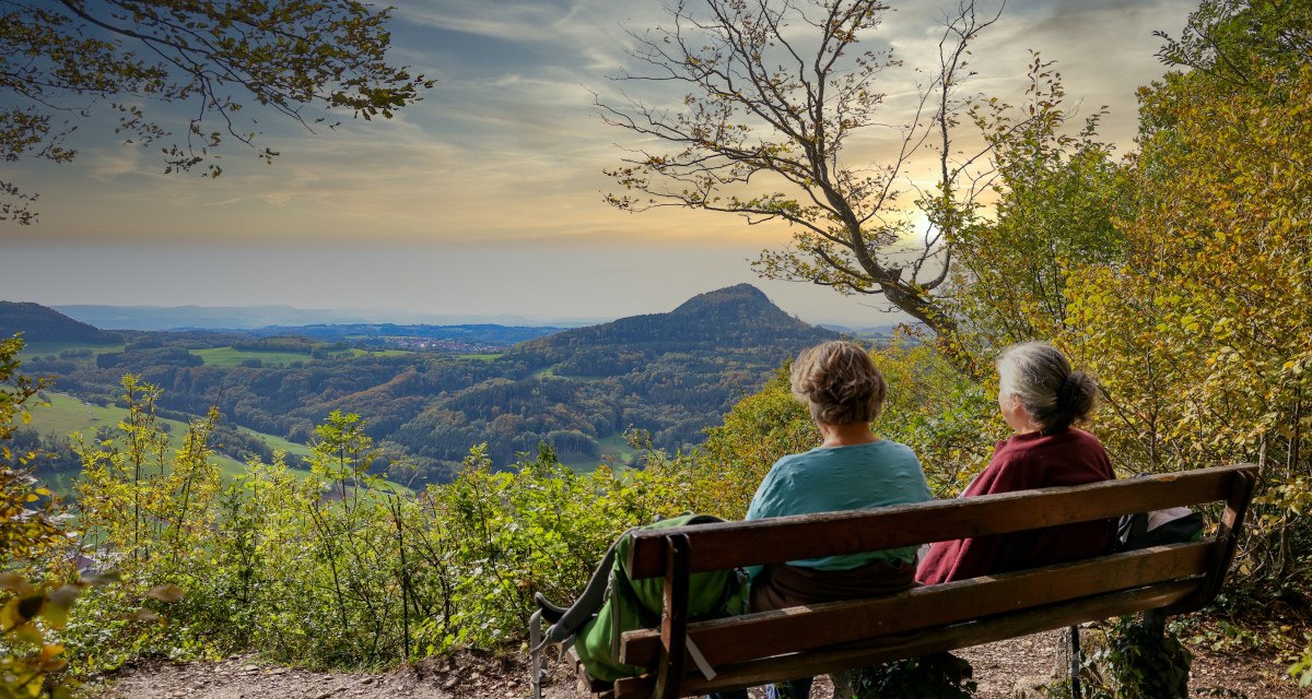 Zwei Personen sitzen auf einer Bank am Aussichtspunkt Luginsland und genießen den Blick auf die hügelige Landschaft im Sonnenuntergang., © Foto: Mario Klaiber