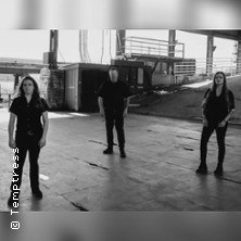 Drei Personen stehen in einem industriellen Umfeld. Die Szene wirkt ruhig und konzentriert, mit Betonboden und metallischen Strukturen im Hintergrund., &copy; links im Bild