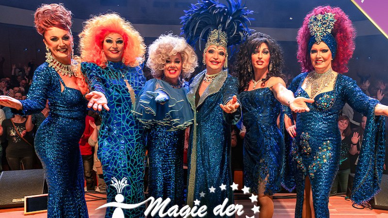 Sechs Drag-Performer in glitzernden blauen Kostümen posieren auf einer Bühne. Im Hintergrund ist ein applaudierendes Publikum zu sehen., © Agentur-Kultur