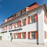 Weißes Gebäude mit roten Fensterläden, blauer Himmel. Schriftzug: 'campuszwei Hotel & Boardinghouse'. Moderne Architektur, sonniger Tag., © TOMAS