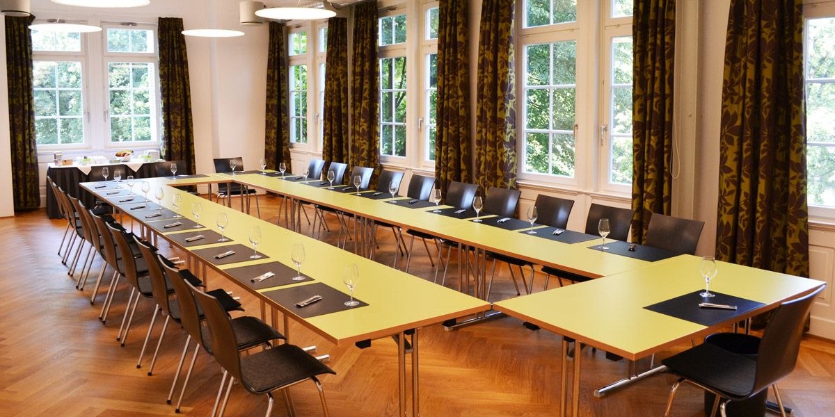Ein heller Tagungsraum mit gelben Tischen in U-Form, schwarzen St&uuml;hlen und gro&szlig;en Fenstern mit Vorh&auml;ngen. Gl&auml;ser und Schreibutensilien sind bereitgestellt., &copy; ECOINN Hotel am Campus