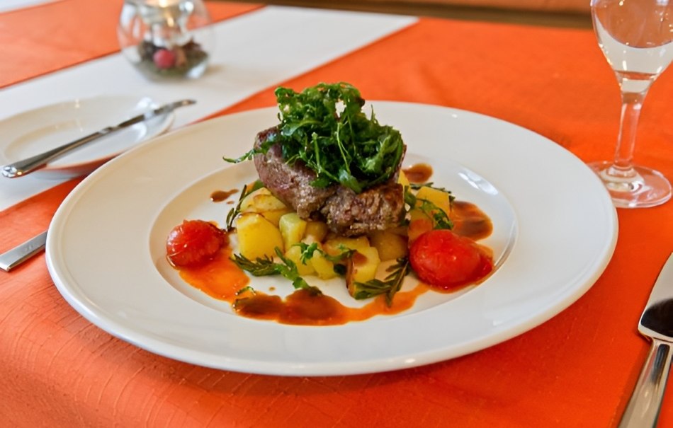 Ein elegantes Gericht mit Fleisch, Kartoffeln, Tomaten und grünem Salat auf einem weißen Teller, serviert auf einem orangefarbenen Tisch., © Hotel Neuwirtshaus Ein elegantes Gericht mit Fleisch, Kartoffeln, Tomaten und grünem Salat auf einem weißen Teller, serviert auf einem orangefarbenen Tisch., © Hotel Neuwirtshaus