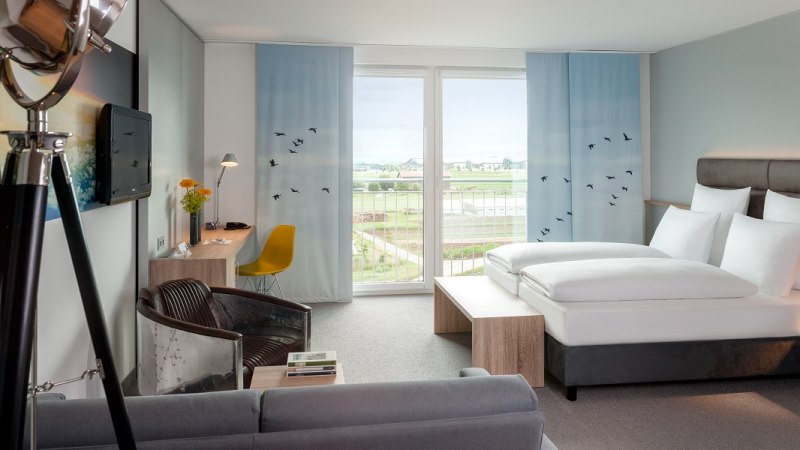 Modernes Hotelzimmer mit gro&szlig;em Bett, Schreibtisch, Sessel und Balkon mit Aussicht auf Landschaft. Dekor mit blauen Vorh&auml;ngen und Vogelmotiven., &copy; Essential by Dorint
