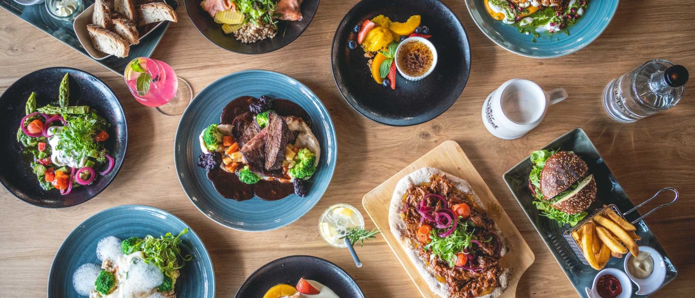 Ein Tisch mit verschiedenen Gerichten: Fleisch mit Gemüse, Salate, Burger mit Pommes, Brot und Desserts. Getränke sind ebenfalls zu sehen., © SatzDrei