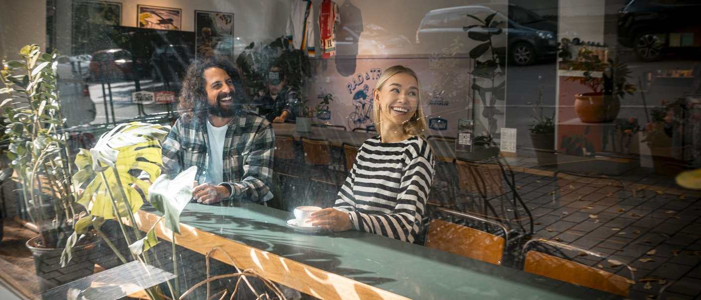 Zwei Personen sitzen l&auml;chelnd in einem Caf&eacute;, umgeben von Pflanzen. Sie genie&szlig;en eine entspannte Atmosph&auml;re, w&auml;hrend sie durch eine Glasscheibe betrachtet werden., &copy; SMG, Sarah Schmid