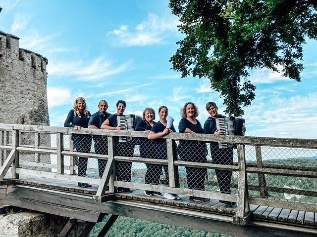 Sechs Personen mit Akkordeons stehen auf einer Holzbr&uuml;cke vor einer Burgmauer, umgeben von B&auml;umen und blauem Himmel., &copy; ConPassione