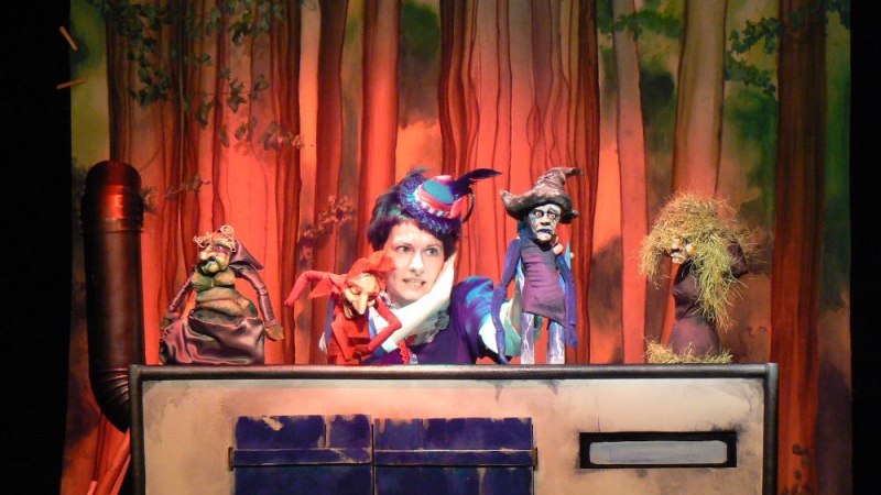 Eine Puppenspielerin mit buntem Hut führt Puppen vor einem Waldhintergrund auf. Verschiedene Charaktere sind auf der Bühne zu sehen., © Ulrike Langenbein Eine Puppenspielerin mit buntem Hut führt Puppen vor einem Waldhintergrund auf. Verschiedene Charaktere sind auf der Bühne zu sehen., © Ulrike Langenbein