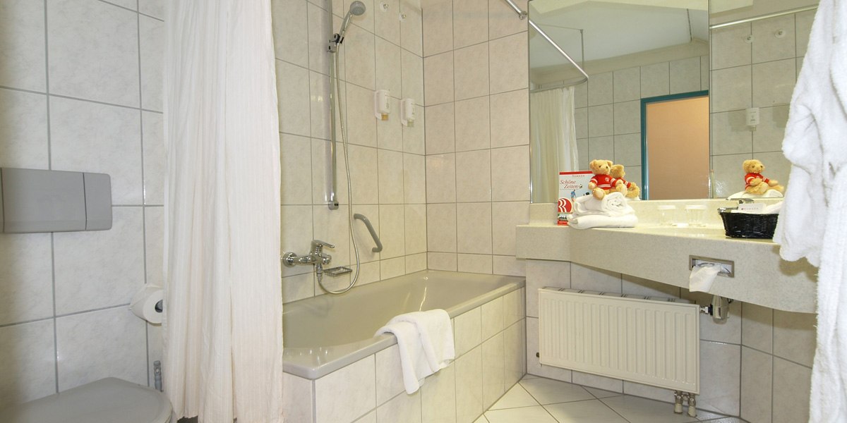 Ein helles Badezimmer mit Badewanne, Duschvorhang, Waschbecken und Spiegel. Handtücher und Dekorationen auf dem Waschbecken., © © H-Hotels.com