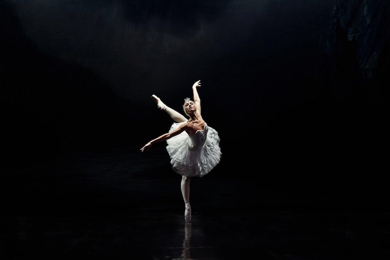 Eine Ballerina in einem weißen Tutu tanzt anmutig auf einer dunklen Bühne, beleuchtet von einem sanften Licht., © Stadthalle Leonberg