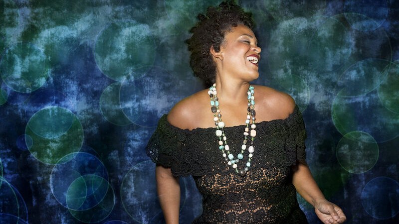 Frau in schwarzem Spitzenkleid und bunter Halskette lächelt vor einem blau-grünen, abstrakten Hintergrund. Sie wirkt fröhlich und entspannt., © Jazzclub Schwäbisch Hall e.V. Frau in schwarzem Spitzenkleid und bunter Halskette lächelt vor einem blau-grünen, abstrakten Hintergrund. Sie wirkt fröhlich und entspannt., © Jazzclub Schwäbisch Hall e.V.