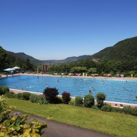 Ein Freibad inmitten einer gr&uuml;nen, bergigen Landschaft. Menschen schwimmen im gro&szlig;en Becken, umgeben von B&auml;umen und H&uuml;geln unter klarem Himmel.