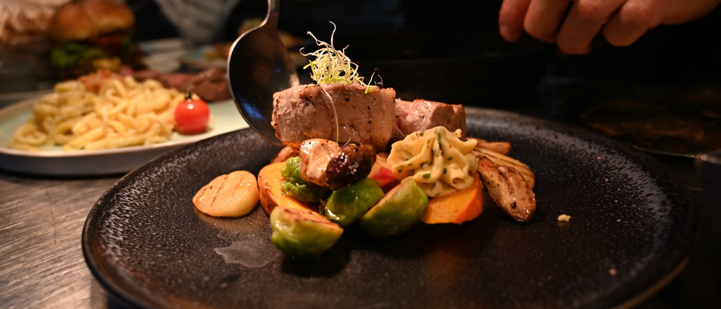 Ein Koch richtet ein Gericht mit Fleisch, Gemüse und Beilagen auf einem schwarzen Teller an. Im Hintergrund sind weitere Speisen zu sehen., © Bad Urach Tourismus Ein Koch richtet ein Gericht mit Fleisch, Gemüse und Beilagen auf einem schwarzen Teller an. Im Hintergrund sind weitere Speisen zu sehen., © Bad Urach Tourismus