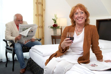 Älteres Paar in einem Hotelzimmer. Frau sitzt auf dem Bett mit Wein, Mann liest Zeitung. Gemütliche Atmosphäre., © Jeanette Bender Älteres Paar in einem Hotelzimmer. Frau sitzt auf dem Bett mit Wein, Mann liest Zeitung. Gemütliche Atmosphäre., © Jeanette Bender