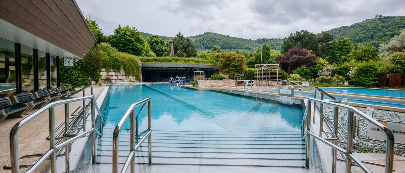 Ein Au&szlig;enpool in einer Therme, umgeben von &uuml;ppiger Vegetation und H&uuml;geln im Hintergrund. Metallgel&auml;nder f&uuml;hren ins Wasser., &copy; Thomas Niederm&uuml;ller