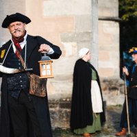 Person in historischer Kleidung mit Laterne und Hellebarde, zwei weitere Personen im Hintergrund vor einem alten Gebäude., © Stadt Nürtingen