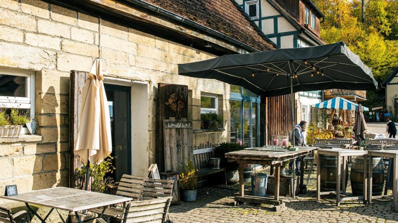 Gemütlicher Café-Außenbereich mit Holzmöbeln und Sonnenschirm, umgeben von Fachwerkhäusern und herbstlicher Landschaft., © Stuttgart-Marketing GmbH, Sarah Schmid Gemütlicher Café-Außenbereich mit Holzmöbeln und Sonnenschirm, umgeben von Fachwerkhäusern und herbstlicher Landschaft., © Stuttgart-Marketing GmbH, Sarah Schmid